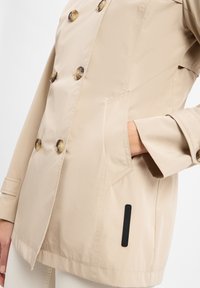 Beige Hochleistungsjacke mit doppelter Knopfleiste aus glattem Stoff, mit großen Knöpfen, einer Seitentasche und einem schwarzen Labelakzent nahe dem Saum.