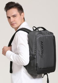Sac à dos gris au design épuré, fabriqué à partir de matériaux durables. Il dispose de plusieurs zippers et d'un logo de marque bien en vue à l'avant.