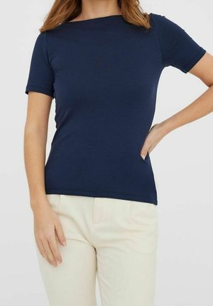 Femme portant une chemise bleu marine ajustée à manches courtes et un pantalon crème, une main sur la hanche, devant un fond clair uni.