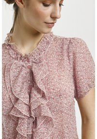 Donna che indossa una blusa floreale rosa e bianca con colletto a volant e parte frontale arricciata, dotata di piccoli bottoni e maniche a sbuffo corte.