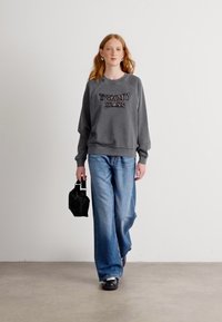 Felpa grigia con logo ricamato "TOMMY JEANS", abbinata a jeans a gamba larga blu. Il modello tiene una piccola borsa nera. Sfondo semplice.