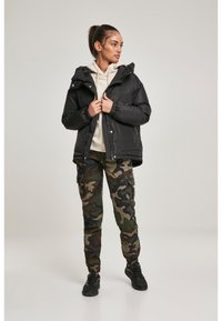 Juoda pūkinė striukė su gaubtu, lengvos konstrukcijos, su užtrauktuku priekyje ir elastiniais rankogaliais. Derinama su kamufliažo krovinių kelnėmis ir juodais sportbačiais.