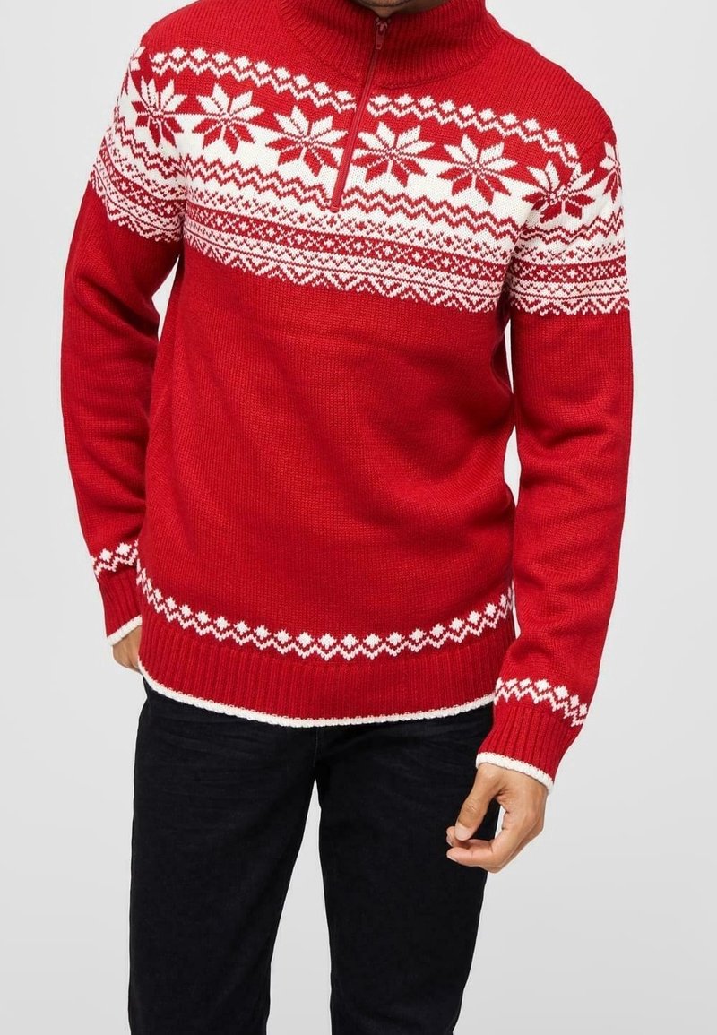 Homme portant un pull rouge en tricot avec des motifs de flocons de neige blancs et géométriques, col zippé, et pantalon noir, debout devant un fond uni.