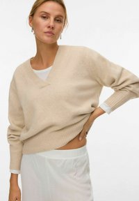 Beżowy sweter dzianinowy z dekoltem w serek i ściągaczami. Ma długie rękawy i luźny krój, w zestawieniu z białą, fakturowaną spódnicą.