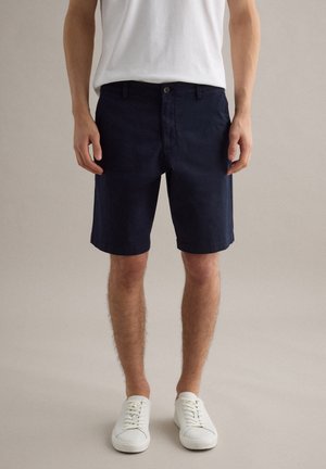 Mann trägt marineblaue knielange Shorts, weißes T-Shirt und weiße Sneakers und steht vor einem einfarbigen, hellen Hintergrund.