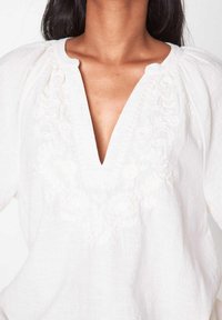 Witte linnen top met een V-hals, met geborduurde bloemendetails en gathered accenten bij de halslijn, zachte textuur en een losse pasvorm.