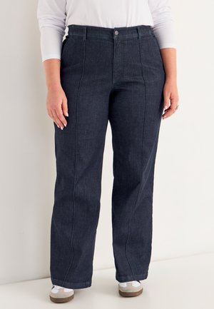 Persoon in donkerblauwe rechte jeans, wit langgerm shirt en witte sneakers, staand tegen een effen achtergrond.