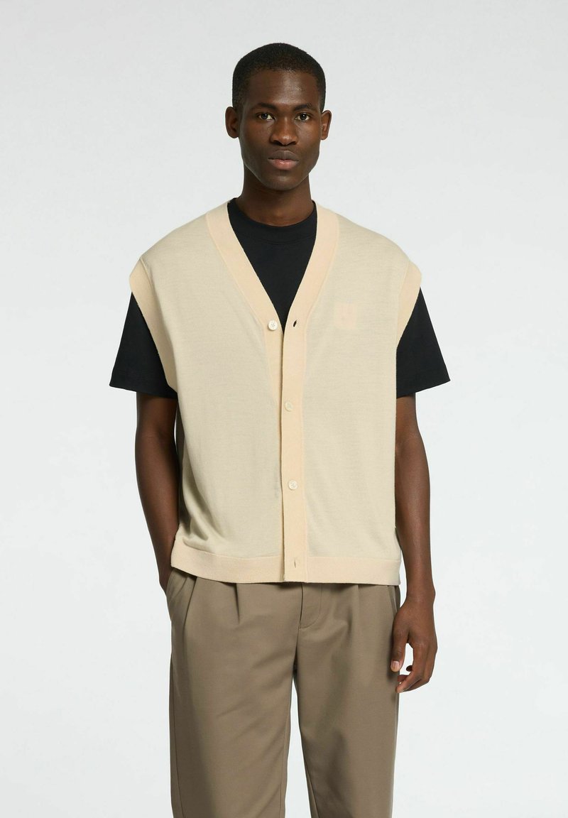 Bež brezrokavnik s globokim V-izrezom, z gumbnim zapiranjem in rebrastim robom, oblečen preko črne majice. Gladko pleteno teksturo.