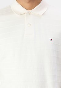 Kermanvärinen polo-paita, jossa on hienovaraiset pystysuorat raidat, lyhyet hihat, kauluspää ja kaksi nappia. Pieni logo vasemmassa rinnassa.