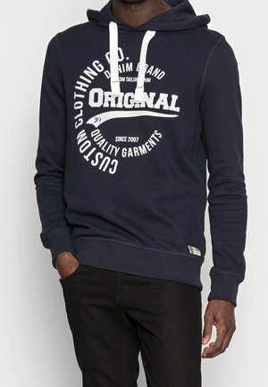 Homme portant un sweat à capuche bleu marine avec des cordons blancs et un texte blanc indiquant "Original Custom Clothing Co Quality Garments."