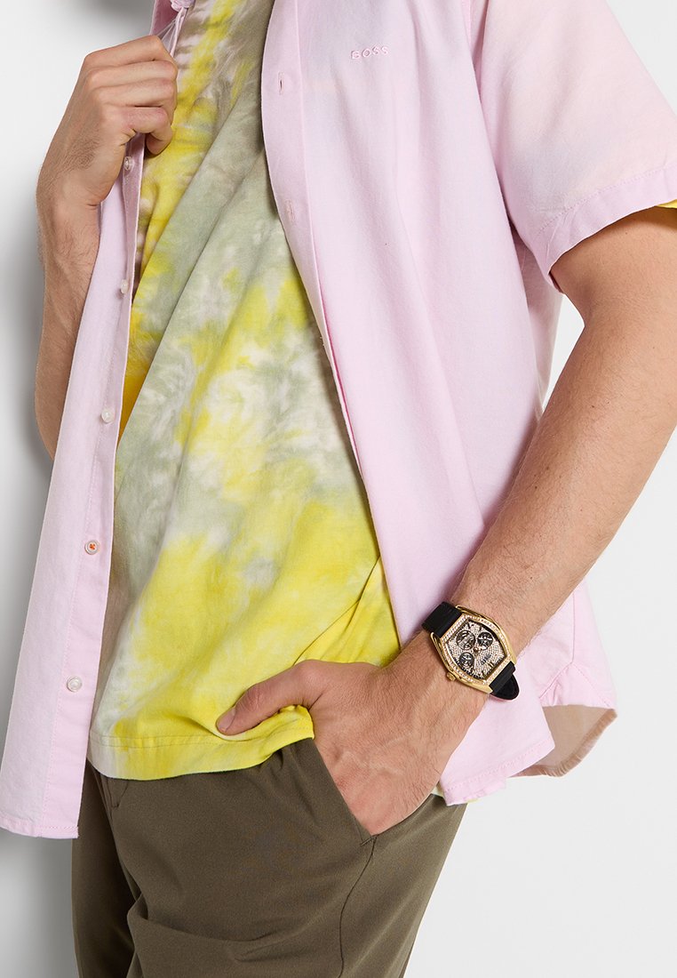 Camisa rosa claro de manga corta superpuesta sobre una camiseta amarilla con efecto tie-dye; pantalones verde olivo oscuros; reloj dorado con correa negra en la muñeca.