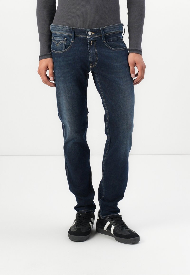 Personne portant un jean slim bleu foncé et des baskets noires avec des bandes blanches, debout devant un fond blanc uni.