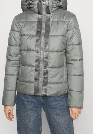 Person trägt eine hellgraue Steppjacke mit glänzendem vertikalem Reißverschlussbesatz und blaue Denim-Jeans vor einfachem Hintergrund.