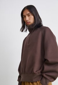 Brun zip-up sweatshirt med hög krage, ribbade ärmmuddar och nederkant. Mjuk textur, lös passform, med en logga på vänster ärm.