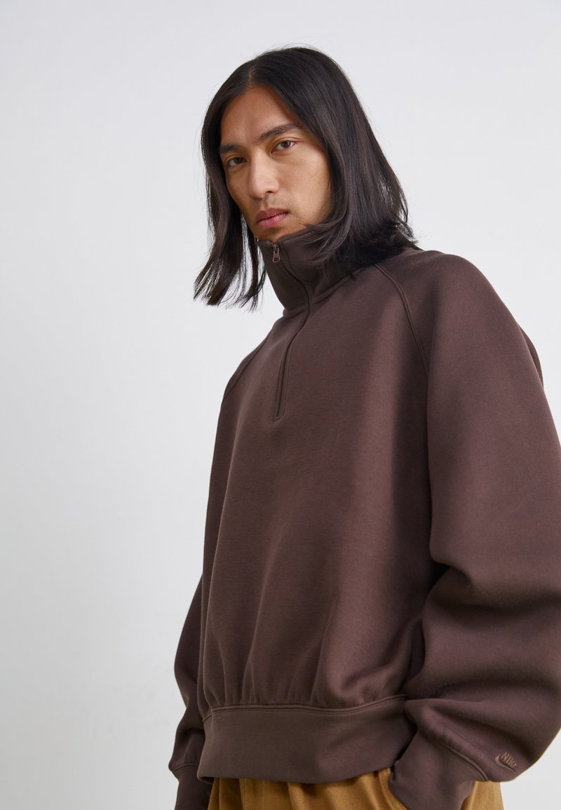 Sudadera marrón con cremallera y cuello alto, puños y dobladillo acanalados. Textura suave, corte holgado, con un logotipo en la manga izquierda.