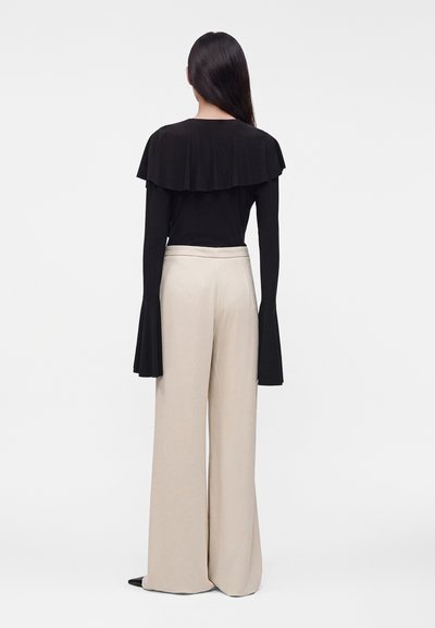 Femme aux longs cheveux foncés portant un haut noir avec un large col à volants et des manches évasées, associée à un pantalon beige large taille haute.
