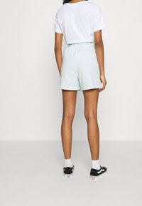 Světle zelené bavlněné šortky s elastickým pasem, kombinované s bílým tričkem a černými teniskami, pohled zezadu.