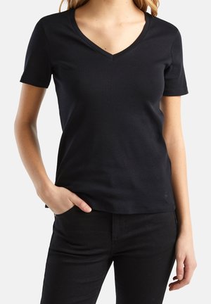 Nainen, jolla on yllään yksinkertainen musta lyhythihainen V-kaula-aukkoinen t-paita, toinen käsi taskussa ja mustat housut, valkoista taustaa vasten.