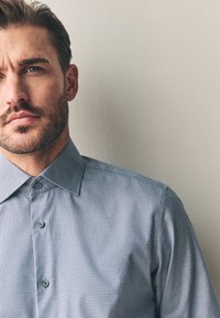 Világoskék kockás ing, klasszikus gallérral, szürke gombokkal és sima szövettextúrával. A háttér semleges.