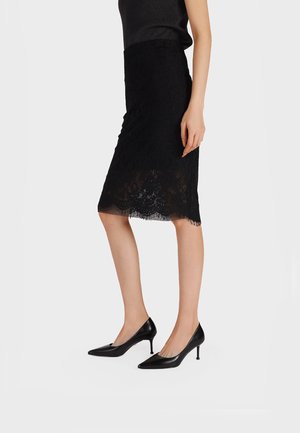 Donna che indossa una gonna nera in pizzo fino al ginocchio e scarpe nere con tacco medio e punta affilata, in piedi davanti a uno sfondo bianco.