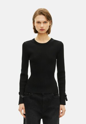 Giovane donna che indossa un top lungo aderente nero a coste con piccoli fiocchi vicino ai polsi e pantaloni neri a vita alta, sfondo neutro.