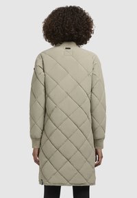Cappotto lungo trapuntato di un beige chiaro con polsini a coste. Presenta un colletto alto e un'etichetta posteriore rettangolare. Tessuto liscio e testurizzato.