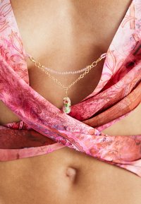 Zlatý vícevrsvený náhrdelník s květinovým přívěskem, krystalovými korálky a jemným řetízkem, na pozadí růžového vzorovaného látkového topu.