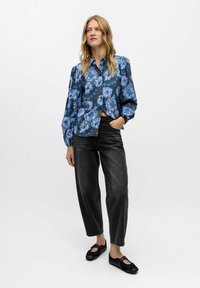 Camicia a fiori con bottoni, in blu e grigio scuro, taglio morbido, maniche a sbuffo. Abbinata a pantaloni in denim neri a vita alta e scarpe nere.