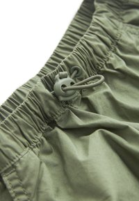 Gros plan d'une ceinture élastique verte avec cordon réglable et verrouillage en plastique sur un vêtement en tissu léger.