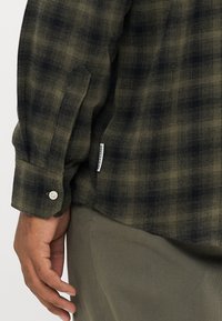 Camicia a maniche lunghe a quadri in verde scuro e nero con polsino con bottone, tessuto testurizzato e una piccola etichetta del marchio sul lato.