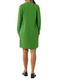 Grün, langärmliges Kleid mit geradem Schnitt, glatter Textur und rundem Ausschnitt. Kombiniert mit schwarzen Schuhen, von hinten betrachtet.