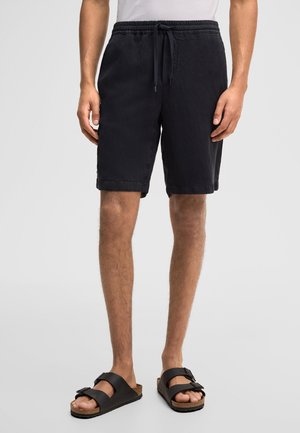 Mężczyzna w czarnych spodenkach ze ściągaczem i czarnych sandałach na klamrę stoi na jednolitym jasnoszarym tle.