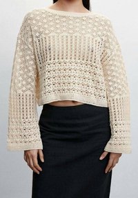 Cremfarbener Häkelpullover mit weiten Ärmeln, der mit texturierten Mustern und einem verkürzten Saum versehen ist, kombiniert mit einem figurbetonten schwarzen Rock.