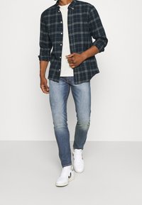 Námořnická kostkovaná košile s límečkem na knoflíky, nošená přes světle šedé tričko, doplněná modrými džínami a bílými teniskami.