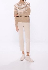 Person, der einen beigen, gemusterten Pullover, helle beigen Hosen und helle beigen Knöchelstiefel trägt, vor einem weißen Hintergrund steht.