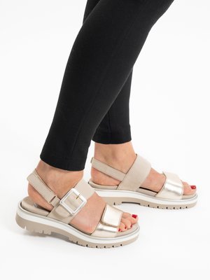 Voeten met beige en metallic open-toe sandalen met een dikke zool en enkelbandjes, gecombineerd met zwarte nauwsluitende broek.