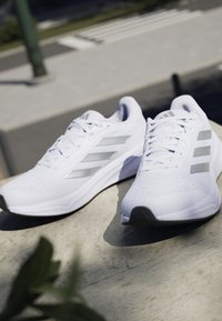 Baskets de sport blanches avec une tige en maille texturée, accent tricolore argenté, et une semelle rembourrée, dotées d'un bout arrondi et d'un laçage.