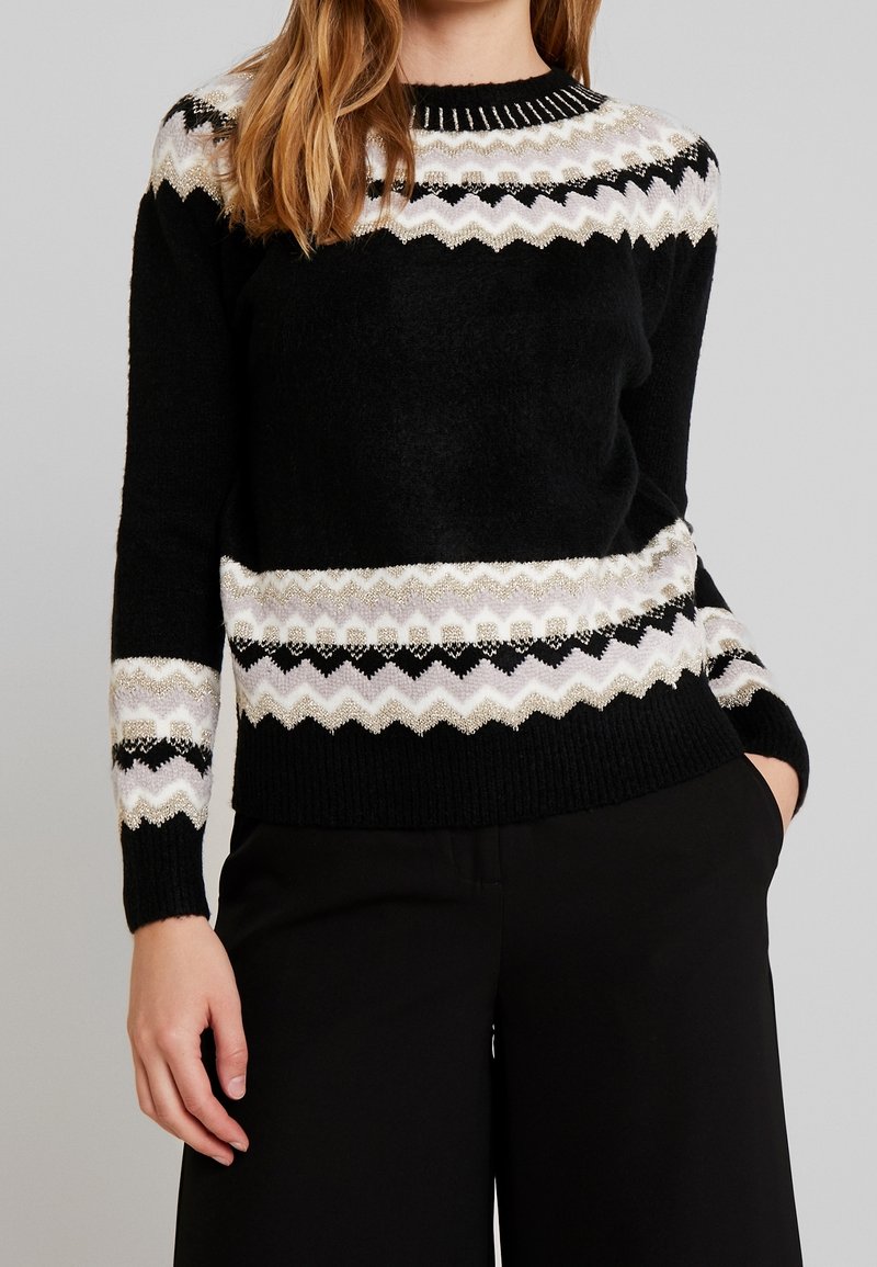 Femme portant un pull noir avec des motifs en zigzag blancs, gris et noirs sur les manches, la poitrine et l'ourlet, associé à un pantalon noir.
