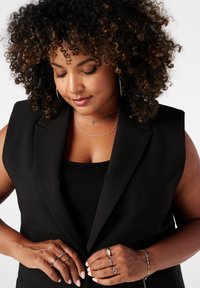 Vrouw met krullend haar die een zwarte mouwloze blazer en top draagt, terwijl ze de blazer met haar handen aanpast en zilveren ringen en armbanden laat zien.