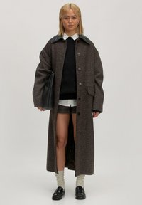 Lange, bruin wollen mixjas met een zwarte leren kraag, knopen aan de voorkant en opgestikte zakken, gecombineerd met een zwarte trui, shorts en zwarte instappers.