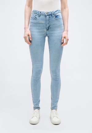 Γυναίκα που φοράει στενά ανοιχτό μπλε τζιν και λευκά αθλητικά παπούτσια, στέκεται μπροστά σε λευκό φόντο.