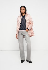 Jachetă pufosă matlasată de culoare roz deschis, cu guler înalt, pulover cu dungi bleumarin dedesubt, pantaloni gri și încălțăminte neagră. Textură netedă, design elegant.