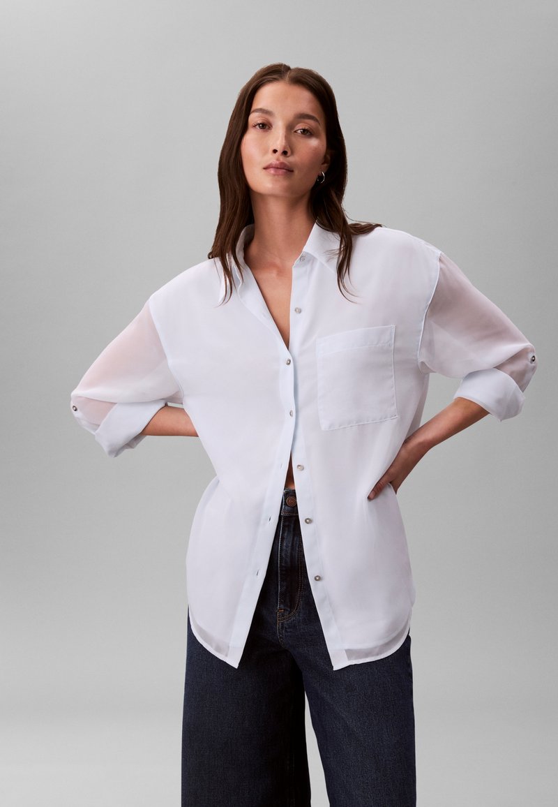 Chemise blanche transparente à boutons, avec une poche poitrine unique, un ourlet arrondi et des manches retrouss ées, associée à un jean foncé taille haute.