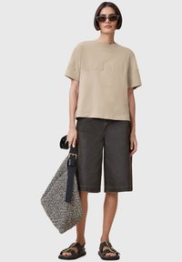 T-shirt in cotone beige con lettering in rilievo, pantaloni bermuda in denim neri e una borsa a fantasia. Occhiali da sole neri e sandali completano il look.