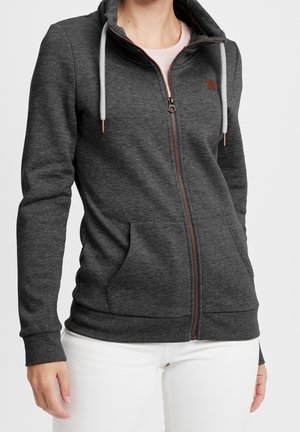 Kobieta ubrana w ciemnoszary, zapinany na zamek sweter z białymi sznurkami i kieszeniami z przodu, na jasnoróżową koszulkę i białe spodnie.