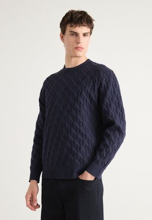 Navy blue knitted sweater featuring a textured pattern, crew neckline, and ribbed cuffs and hem. Worn with dark jeans.