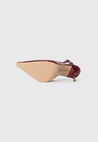 Scarpa stiletto a punta bordeaux con sole beige e fibbia argentata, mostrata dal lato della suola su sfondo bianco.