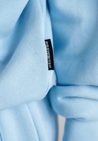 Helles blaues Gewebe mit einer weichen, strukturierten Oberfläche. Ein schwarzes Etikett mit dem Text "VERBOTEN" ist entlang einer Kante sichtbar.