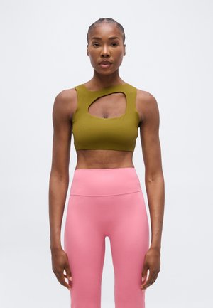 Donna in piedi di fronte, indossa un crop top verde oliva con un'apertura sul petto e leggings rosa a vita alta.
