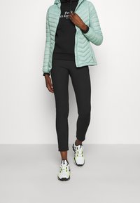 Hellgrüne gesteppte Jacke über einem schwarzen Hoodie, kombiniert mit schwarzen Leggings und weißen Sneakers mit gelben Akzenten.